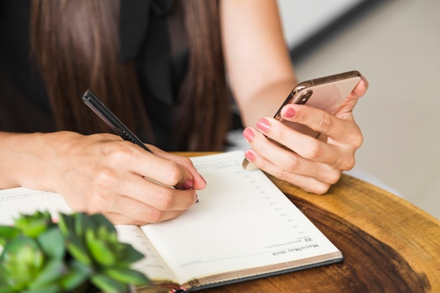 Close-up de mãos de uma mulher escrevendo em um caderno e segurando um smartphone, uma cena que pode envolver a organização ou planejamento de envio de SMS.