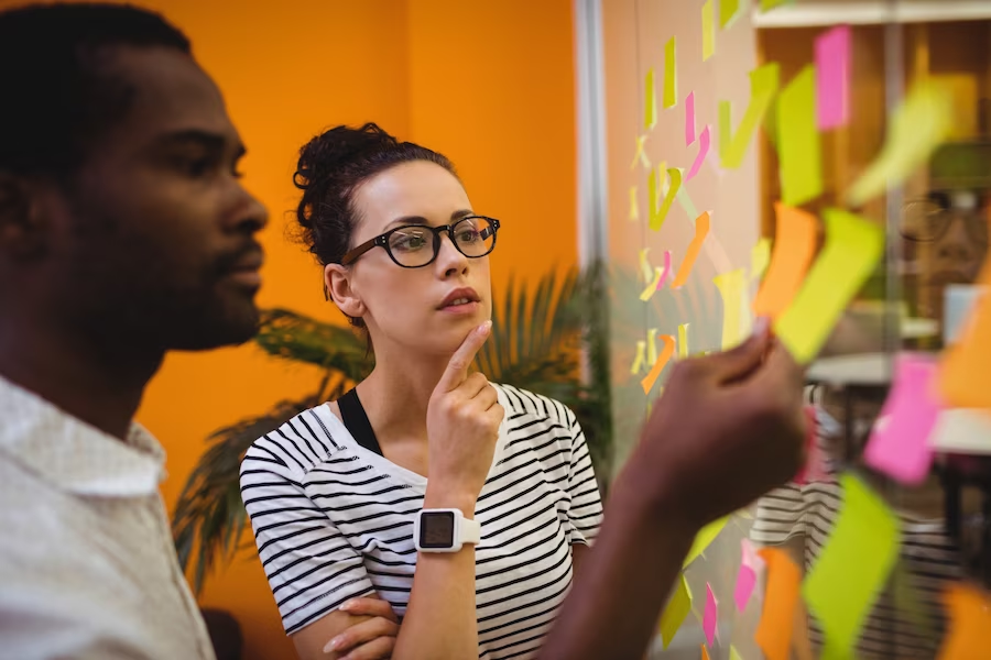 Dois colegas fazendo brainstorming com um clique de compreensão, cercados por post-its coloridos em uma parede de vidro em um escritório criativo.