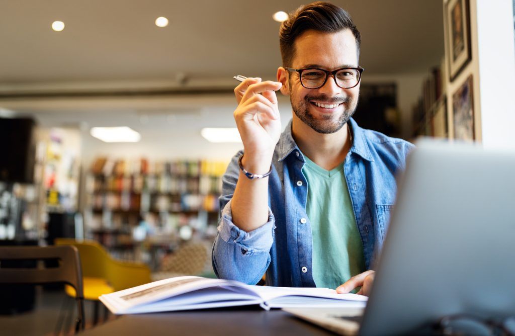 Homem sorridente trabalhando com seu laptop em um ambiente de café, exibindo a satisfação do cliente enquanto realiza uma pesquisa de satisfação online.
