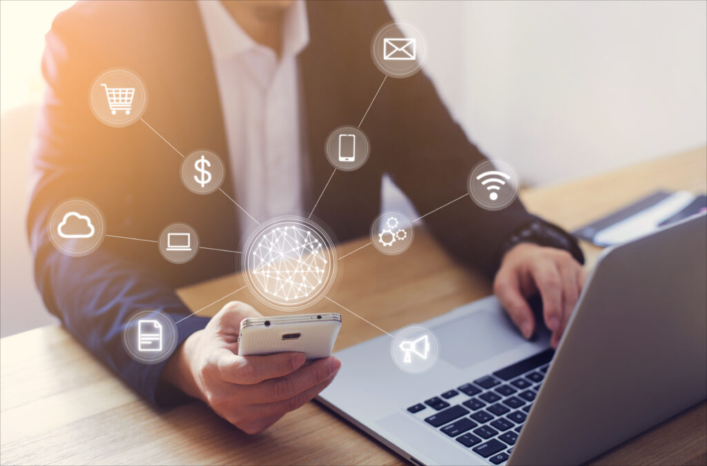 Homem de negócios usando um smartphone enquanto trabalha em um notebook. Ao redor do celular, há ícones digitais representando diferentes canais e serviços — como e-mail, Wi-Fi, nuvem, compras, smartphone, configurações e marketing — conectados a uma rede central, simbolizando integração tecnológica e comunicação omnichannel.