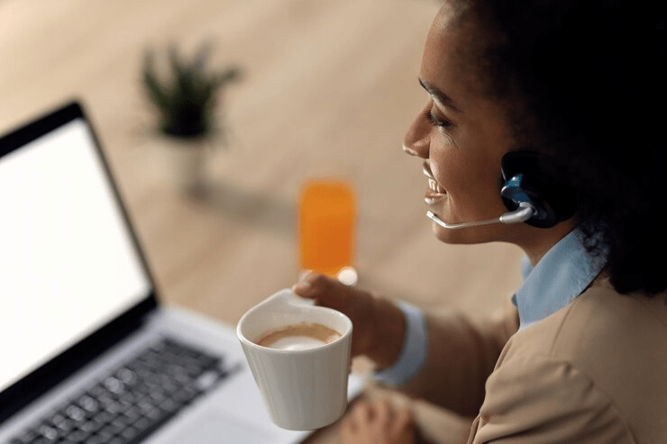 Mulher alegre com headset, segurando uma xícara de café enquanto trabalha no computador, simbolizando multitarefa eficiente e satisfação no trabalho.

