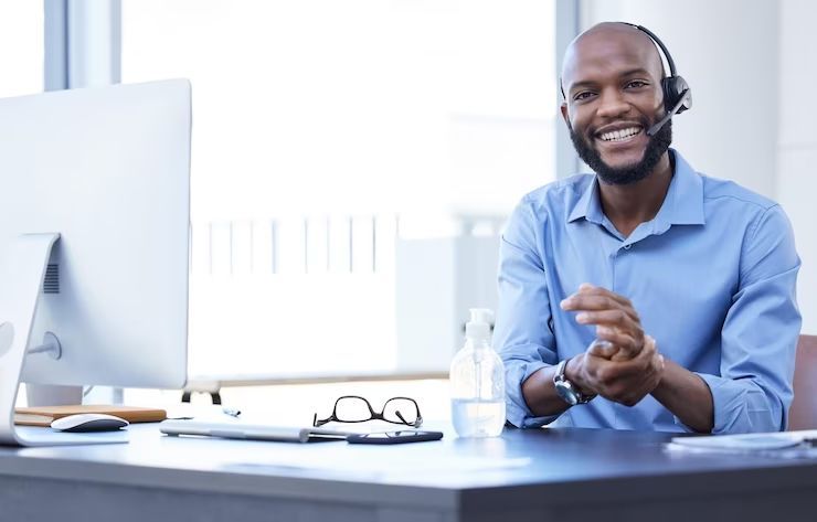 Um homem sorrindo enquanto usa um headset em frente ao computador, ilustrando a adoção de tecnologias de comunicação que podem ajudar a reduzir custos de telefonia.