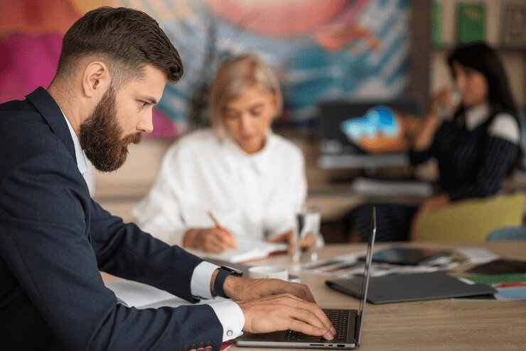Homem concentrado usando um smartwatch e digitando em um laptop em um escritório colaborativo, exemplificando a gestão de tempo e tarefas.

