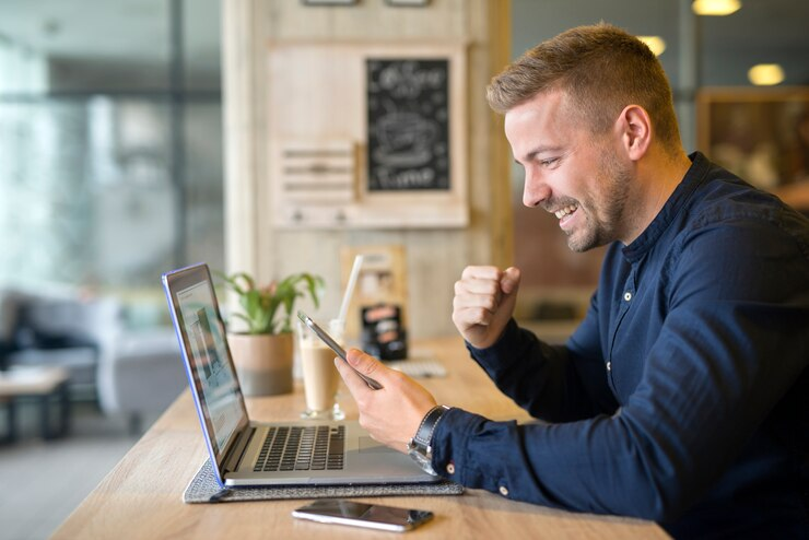 Um homem sorridente em um café trabalha simultaneamente em um laptop e em um smartphone, ilustrando gestão eficiente do tempo e multitarefa.