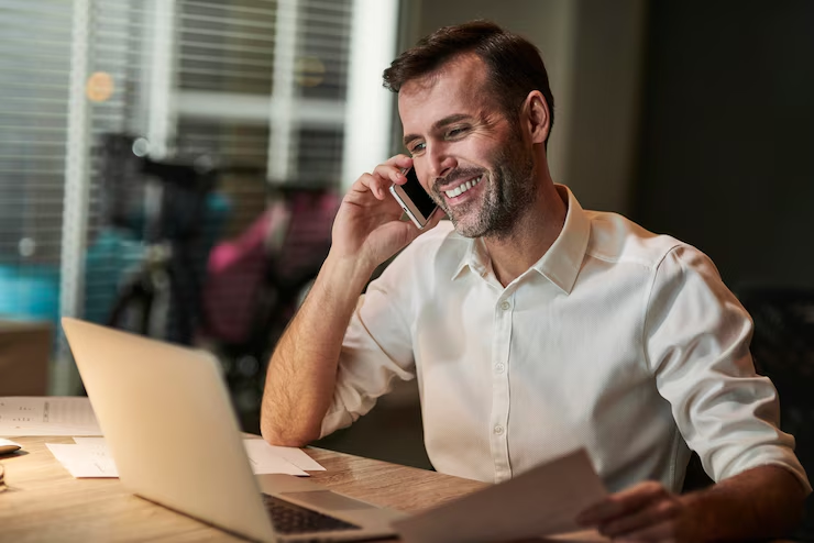 Homem sorridente falando ao telefone, segurando documentos com uma expressão satisfeita, representando a eficiência do serviço 0800 em um ambiente de trabalho.
