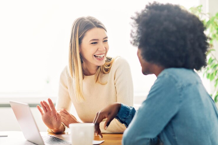 Duas mulheres conversando alegremente em frente a um laptop, uma delas gesticulando animadamente, refletindo a satisfação no pós atendimento.