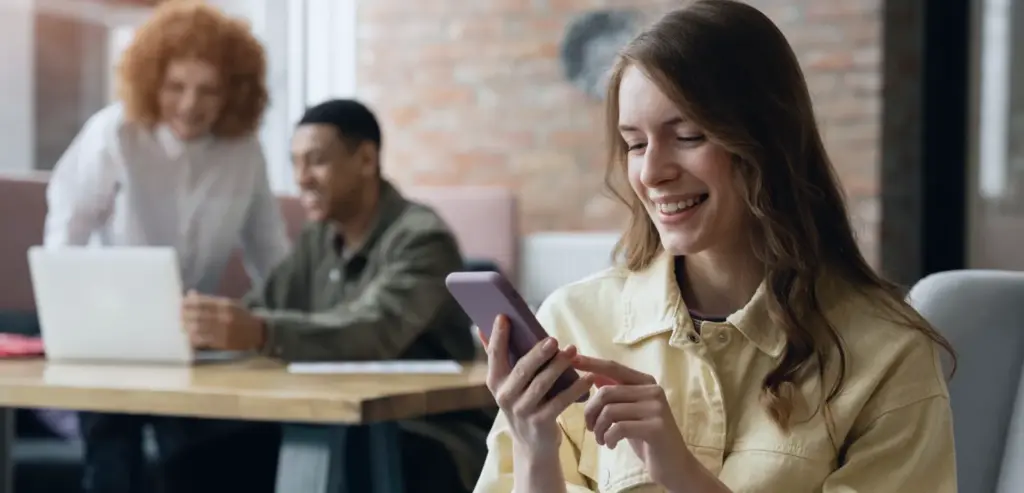 mulher sorrindo enquanto usa o celular em ambiente de trabalho com colegas ao fundo, representando atendimento ao cliente moderno