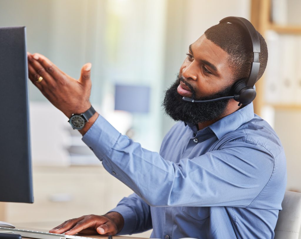  Um homem de camisa azul e com fones de ouvido com microfone está interagindo com uma tela de computador. Ele está gesticulando com a mão aberta em direção à tela, como se estivesse explicando algo do PABX Virtual.