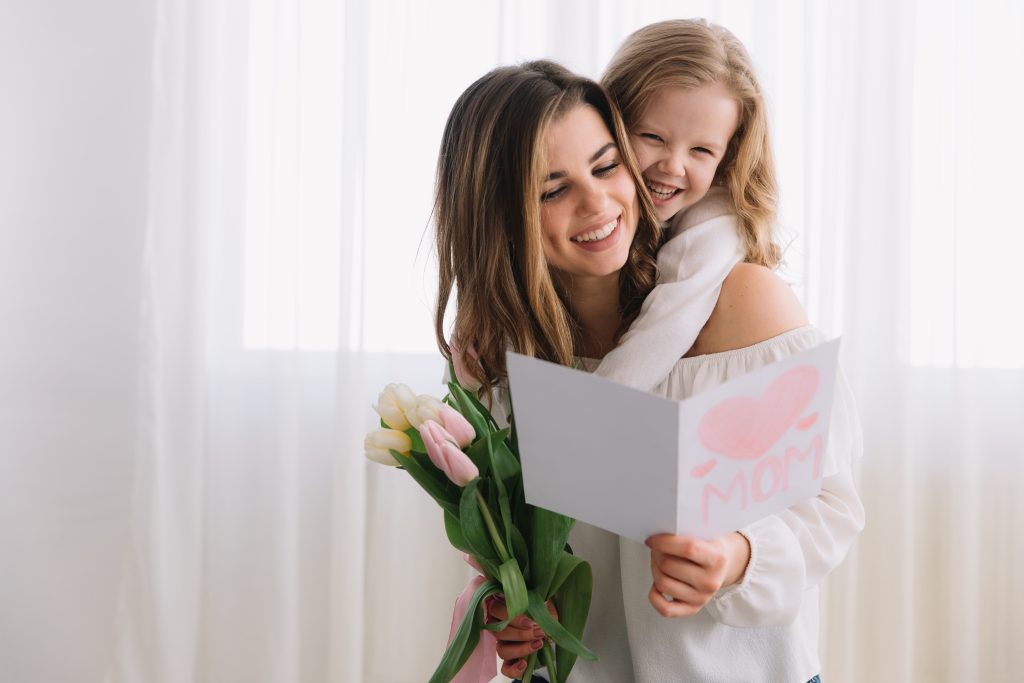 Uma jovem mãe sorridente segurando um buquê de tulipas brancas e rosas, recebendo um cartão com mensagem de Dia das Mães com a palavra "MOM" em um fundo rosa, abraçada por sua filha sorridente em um ambiente claro e arejado.