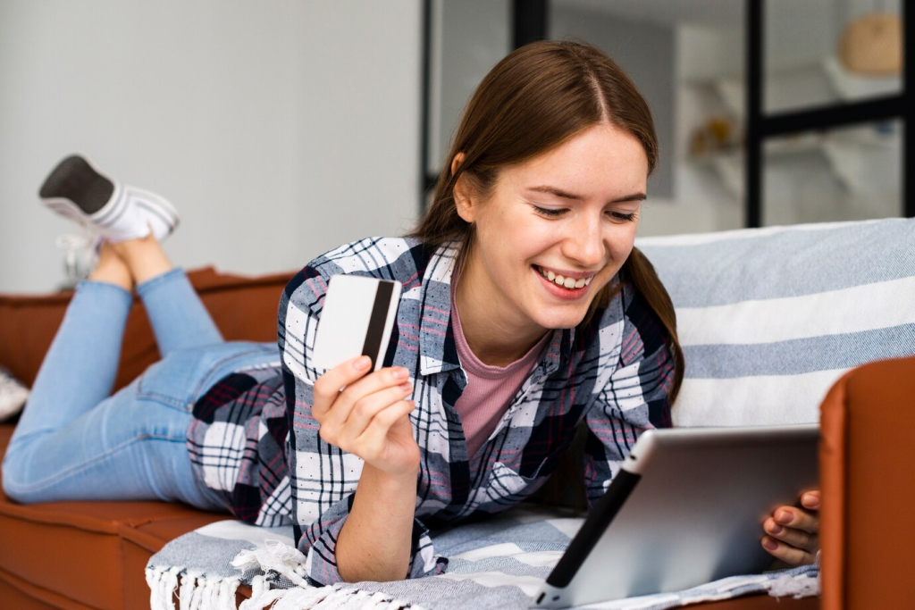 Uma mulher jovem, radiante e descontraída, deitada de bruços em seu sofá, segura um cartão de crédito e olha encantada para seu tablet. É a imagem da comodidade moderna de compras online, onde um simples toque pode trazer alegria instantânea.