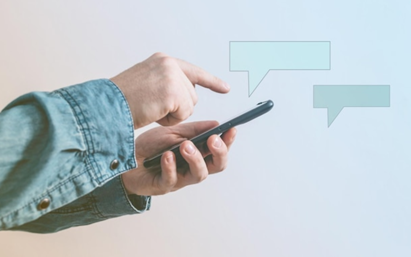 Uma pessoa usando uma jaqueta jeans está digitando em um smartphone com balões de diálogo vazios acima, representando a comunicação digital no atendimento ao cliente através de mensagens de texto ou chat em aplicativos.






