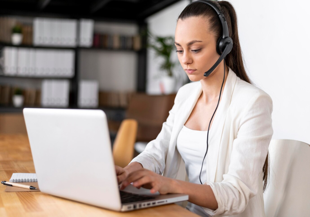 Uma profissional sorridente fala ao telefone enquanto digita em seu laptop, exemplificando o atendimento ao cliente amigável e eficiente de uma secretária remota.
