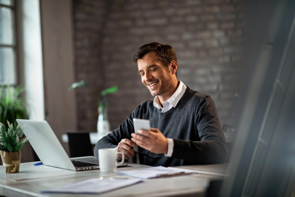Homem sorridente usando um smartphone enquanto trabalha em um laptop num escritório moderno com tijolos à vista.