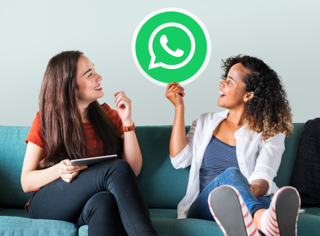 Duas pessoas alegres conversam enquanto uma delas segura um tablet, com um ícone do WhatsApp flutuando entre elas, simbolizando a comunicação fácil e acessível proporcionada pela plataforma.