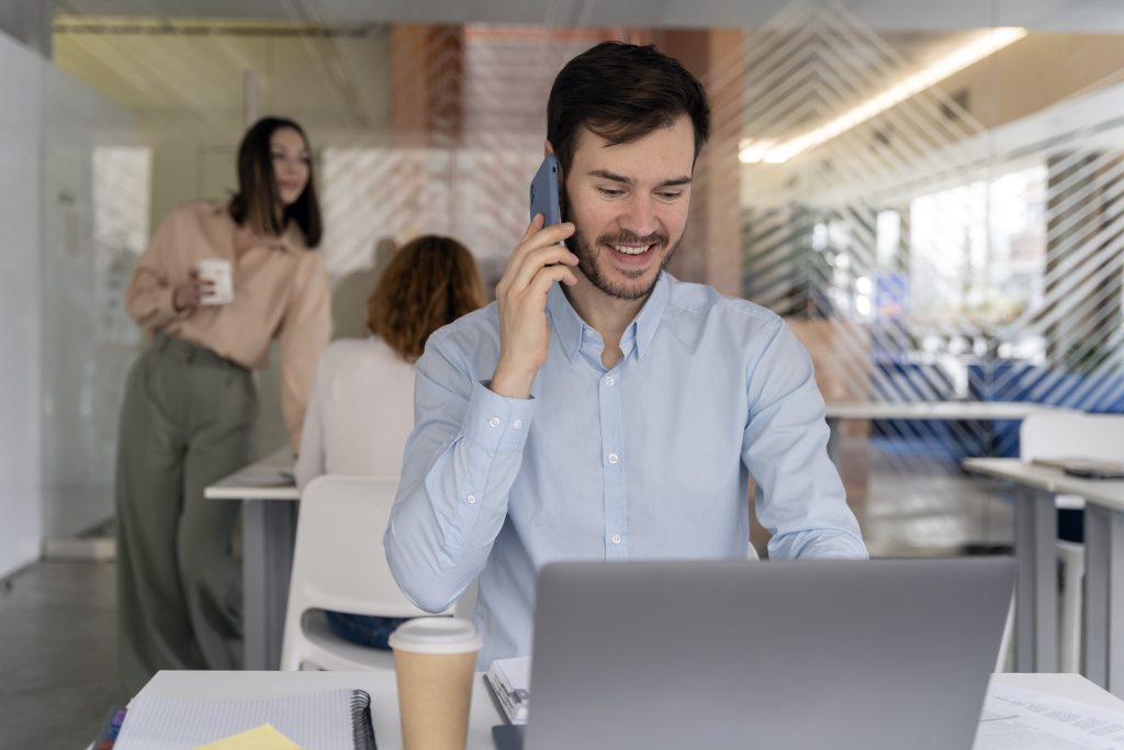 um jovem profissional, sorrindo enquanto fala ao telefone, sentado em sua mesa com um laptop aberto em um escritório moderno e bem iluminado. Ao fundo, seu colega segura uma caneca de café.