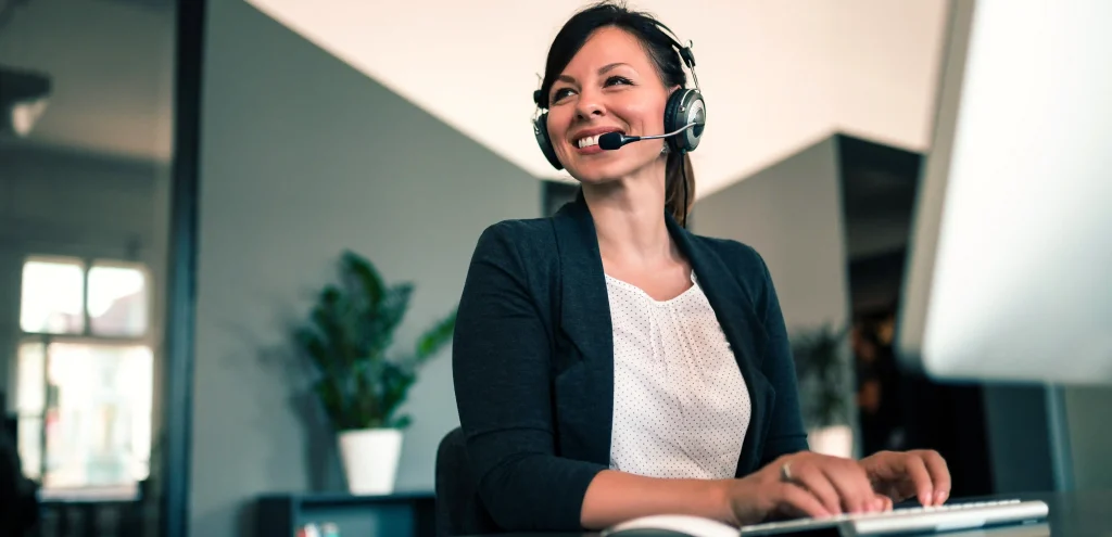 Mulher atendente sorridente com headset, digitando em um computador em ambiente corporativo, representando a terceirização de call center como solução eficiente para atendimento ao cliente.