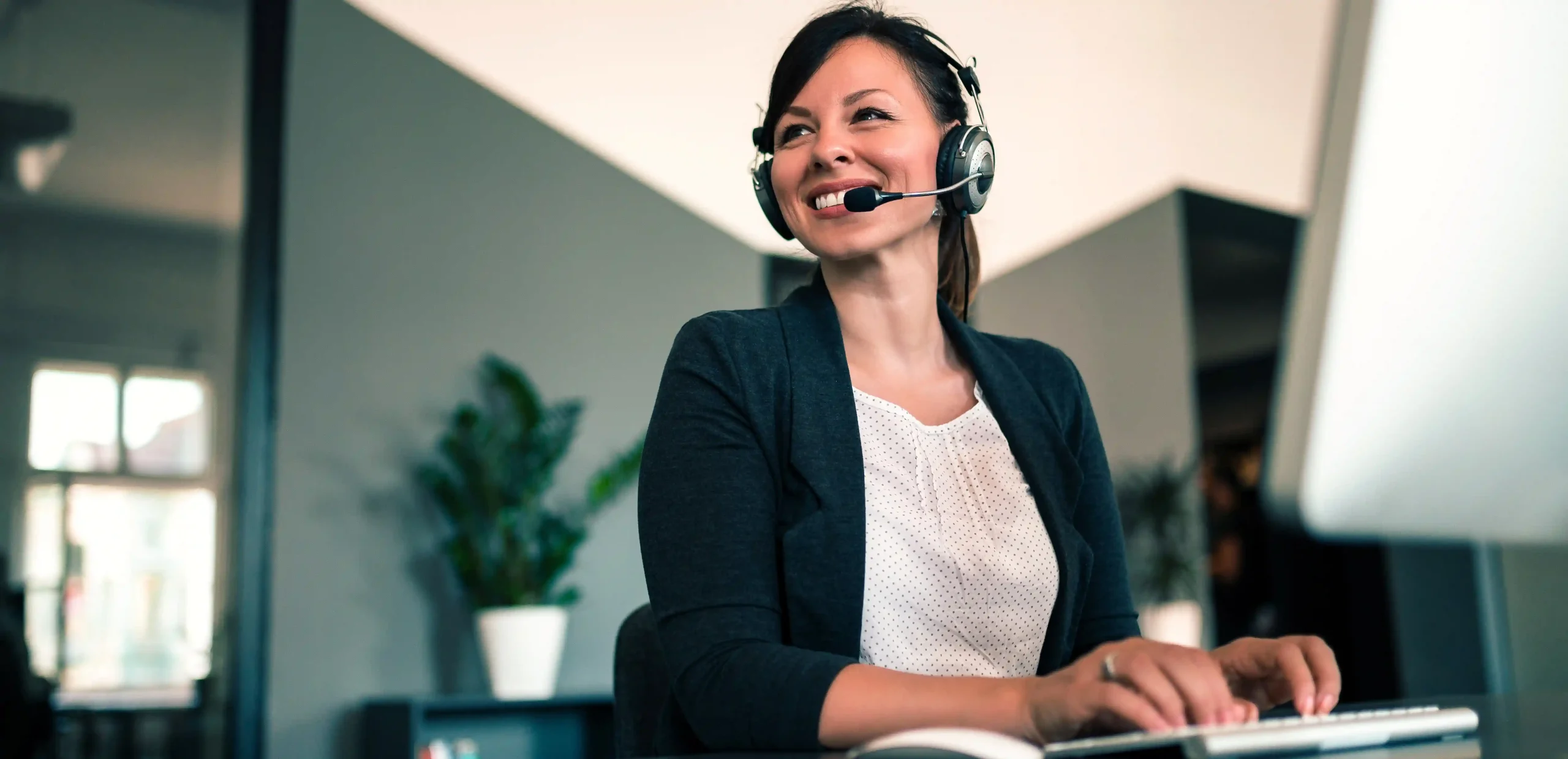 Mulher atendente sorridente com headset, digitando em um computador em ambiente corporativo, representando a terceirização de call center como solução eficiente para atendimento ao cliente.