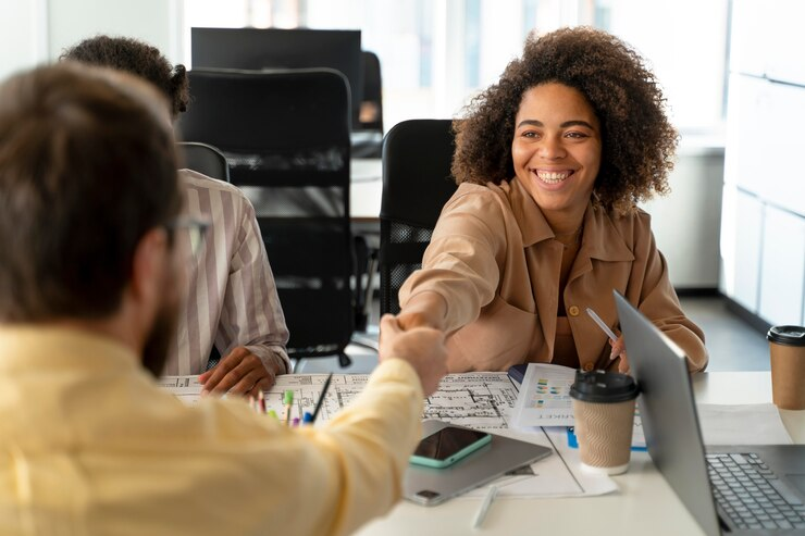 Mulher sorridente em um ambiente de escritório, estendendo a mão para um aperto de mão durante uma reunião, simbolizando um atendimento ao cliente amigável e acolhedor.