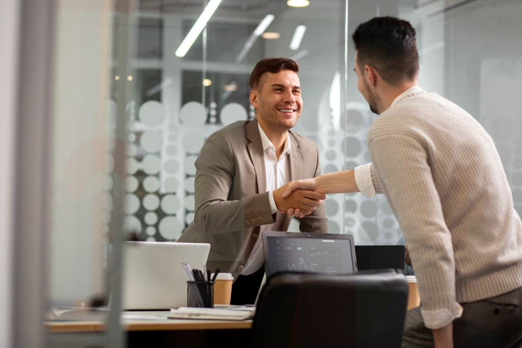 Dois homens de negócios em trajes formais apertando as mãos em um escritório moderno, simbolizando uma parceria ou acordo bem-sucedido.
