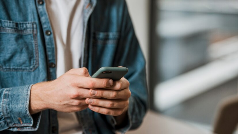 Homem jovem usando um smartphone em um ambiente de trabalho casual, focando em mensagens de texto para comunicação empresarial.