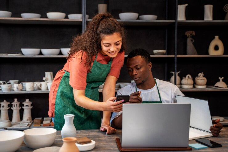 Dois jovens, um homem e uma mulher, usando aventais verdes em uma oficina de cerâmica. Eles estão sorrindo e analisando informações em um smartphone, enquanto o homem também usa um laptop. O ambiente está repleto de cerâmicas e prateleiras ao fundo.