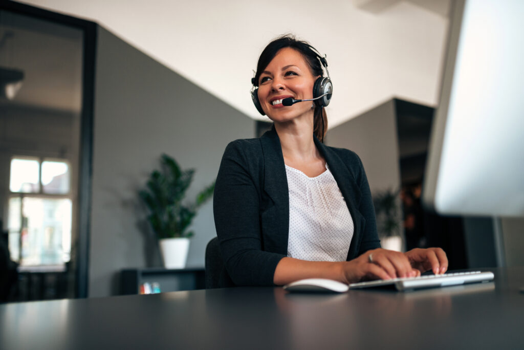 Mulher sorridente com headset atendendo ao cliente em um call center moderno, sentada em frente a um computador, representando um atendimento profissional e eficiente.