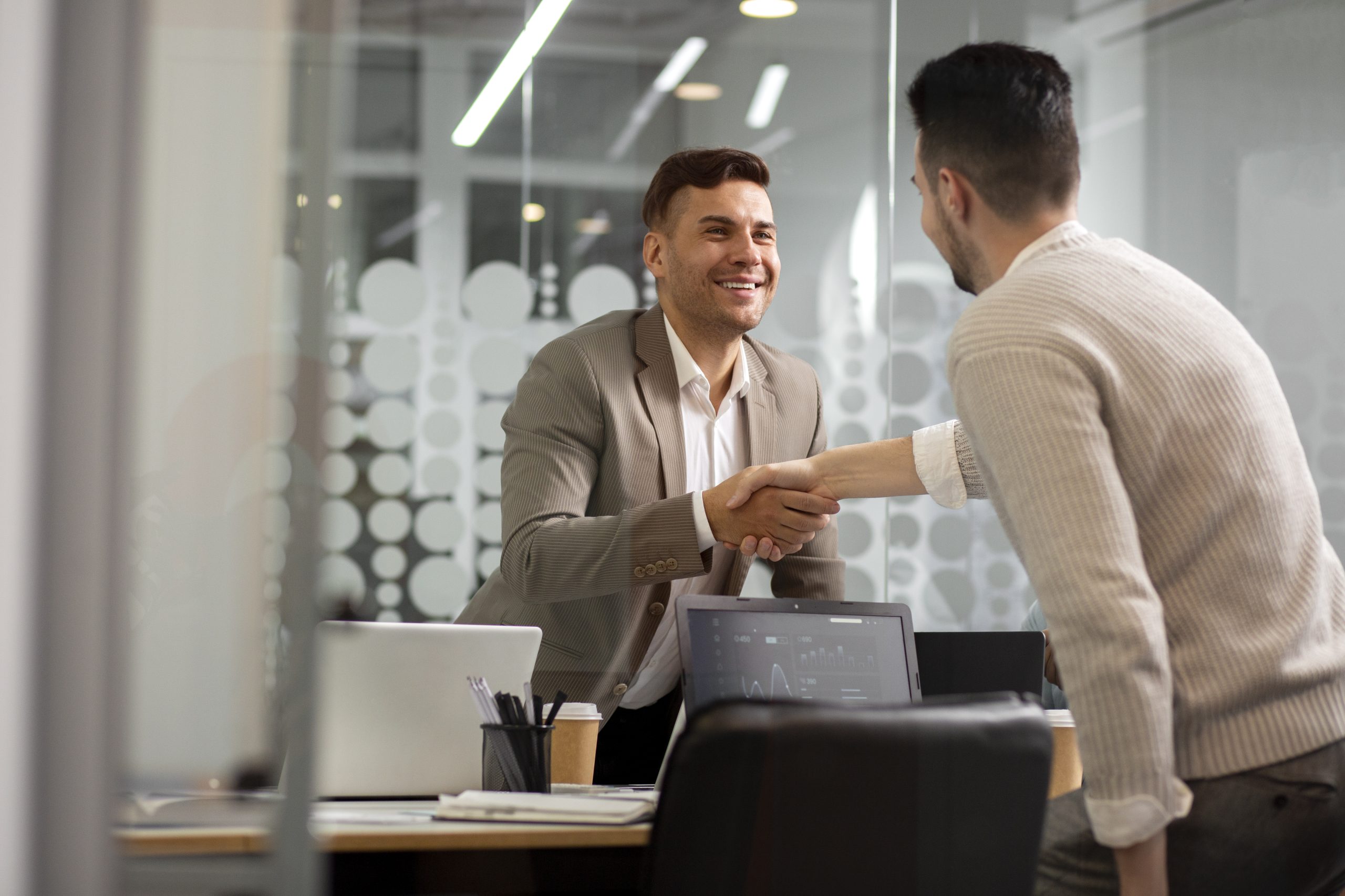 Dois homens apertando as mãos em um ambiente corporativo, sugerindo uma conclusão bem-sucedida de uma reunião de negócios. Um dos homens, em um terno cinza claro, está sorrindo e parece estar satisfeito com o resultado da interação. No fundo, um ambiente de escritório moderno com laptops, canetas e papéis sobre a mesa, e uma parede de vidro com design de bolhas ao fundo, dando um toque contemporâneo ao espaço.