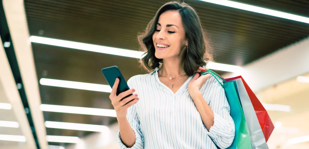 Mulher sorridente segurando sacolas de compras e lendo mensagem no celular, ilustrando uma estratégia eficaz de SMS Marketing para impulsionar vendas.
