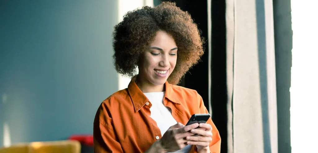 Mulher sorridente usando celular perto da janela, simbolizando praticidade e agilidade com recurso de click-to-call.