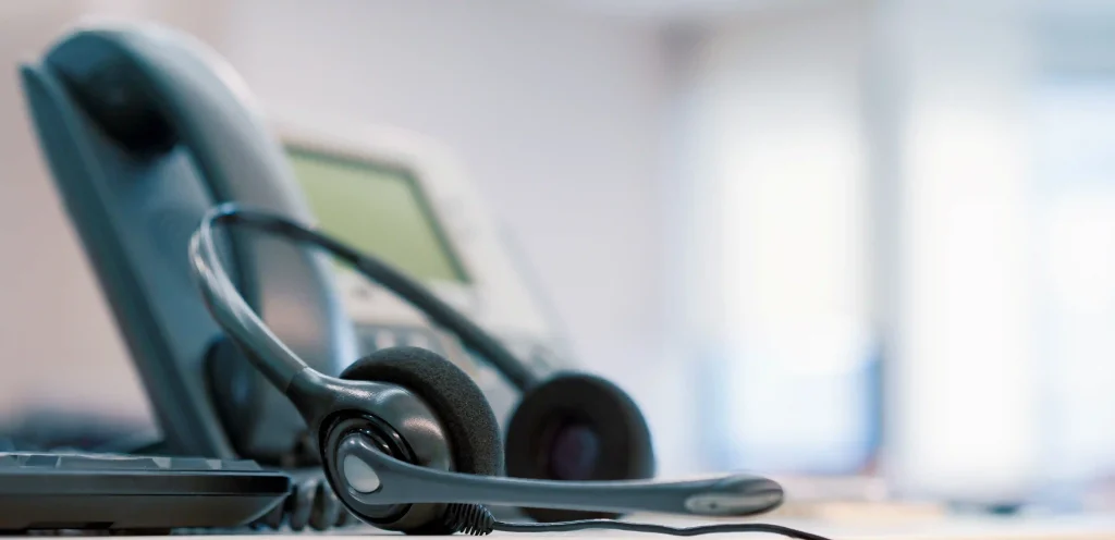 Headset apoiado sobre teclado com telefone ao fundo, representando o início do atendimento automatizado com URA.