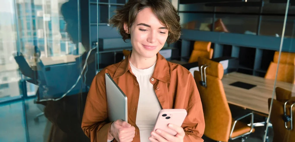 Mulher sorrindo enquanto usa o celular em um ambiente corporativo, representando o uso da API oficial WhatsApp para comunicação empresarial eficiente.