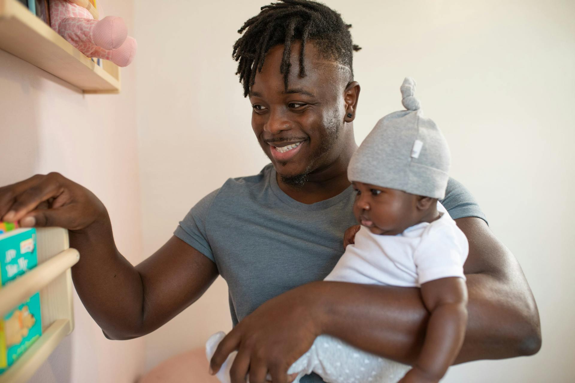 Um pai sorridente segura seu bebê nos braços enquanto alcança uma prateleira. Ele está vestindo uma camiseta azul e o bebê, com um gorro cinza, veste uma roupa branca.