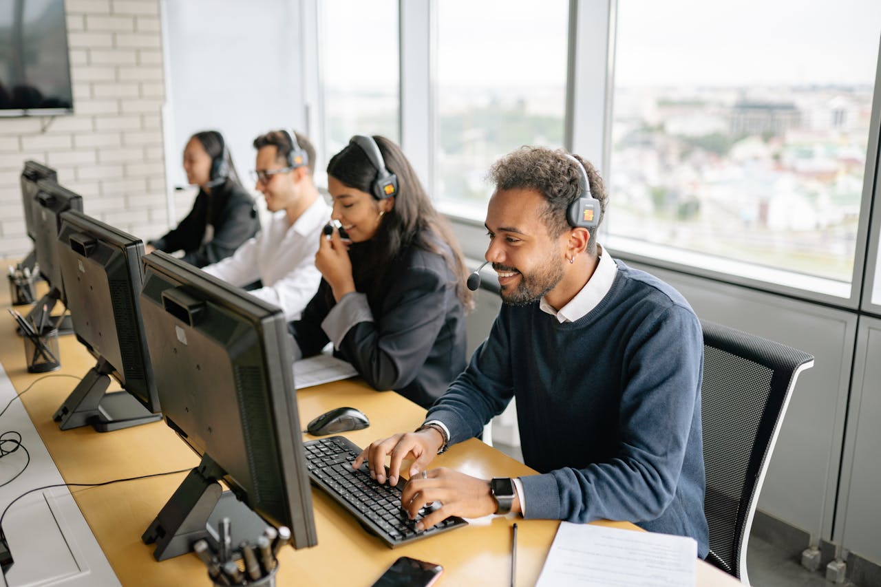 Equipe de atendimento ao cliente em um call center moderno, composta por profissionais usando headsets, trabalhando em computadores. Um dos agentes, um homem de pele morena e barba, está sorrindo enquanto digita no teclado, evidenciando um ambiente de trabalho positivo e colaborativo. As janelas ao fundo oferecem uma vista urbana, indicando que o escritório está localizado em um andar alto de um edifício.