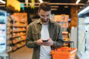 homem jovem com cestinha de compras olhando o celular em supermercado